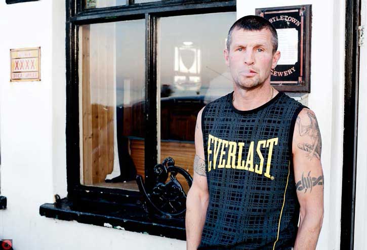 Steve, leaning on the outside wall of a pub, wearing a sleeveless Everlast t-shirt, with tattoos on his arms.