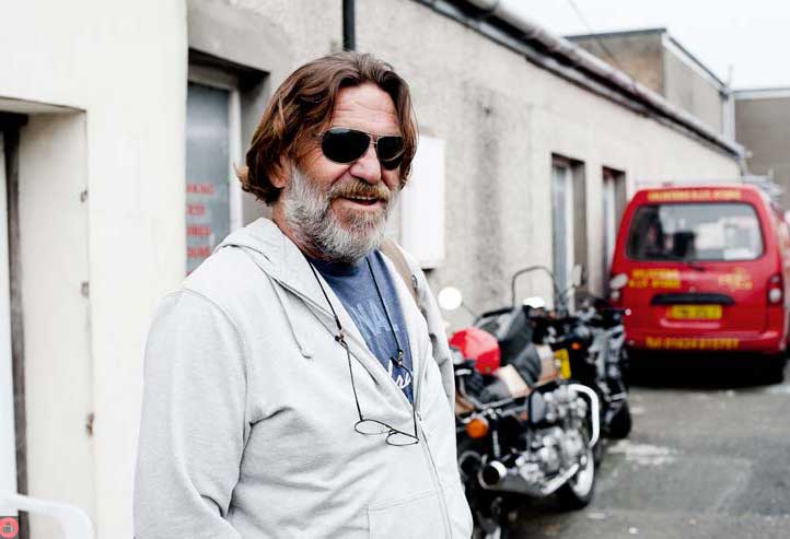 A bearded australian guy, wearing shades, with a Moto Guzzi cruiser motorcycle parked in the background.