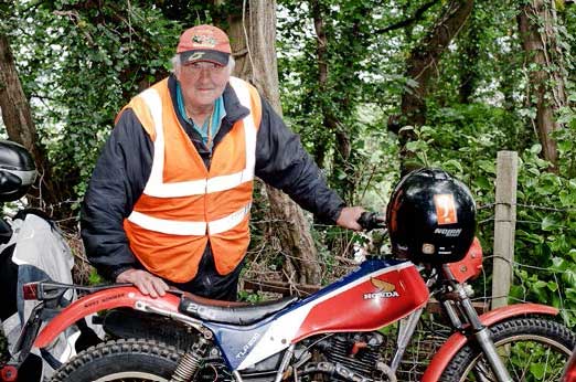 Nippy Norman, a marshal, wearing a day-glo tabard and wearing a baseball cap, leaning on his Honda 250 TLR, looking straight to camera.
