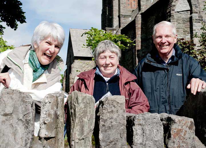 Judy, Joan and John, with a church behind, in the sun, leaning on a wall and grinning and laughing to camera.