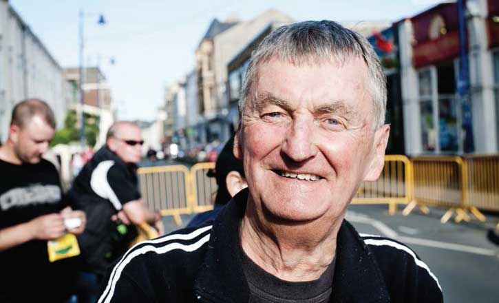 Peter, a local whose lived in Ramsey for 50 years, standing in a street with barriers behind, looking straight to camera with a smile.