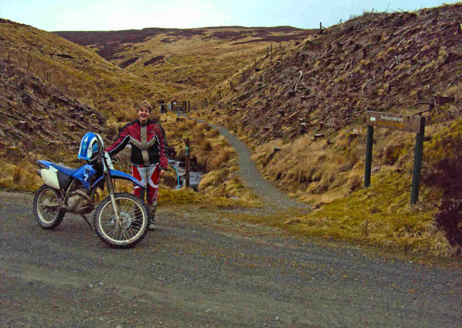 jan having a breather, standing next to the TT-R, with her helmet hanging on the end of the handlebar, on a gravel track.