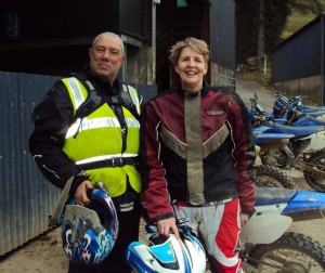 Jan and John, holding their helmets, standing in front of all the bikes, next to a barn.