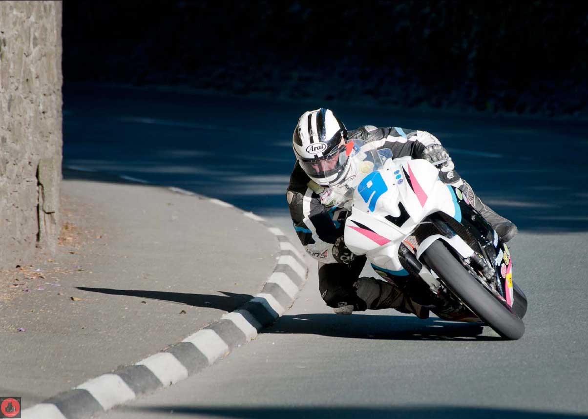 Michael Dunlop on his race bike, cranked over on a right hander, skimming a kerb, with a stone wall edging the pavement.
