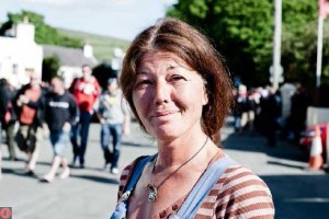 Martina standing in a road wearing a t-shirt and dungarees, with spectators walking past, smiling to camera, 