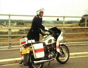 1970's police motorcyclist standing next to his police 650 Triumph Thunderbird wearing an open face helmet, gauntlets and aviator sun glasses.