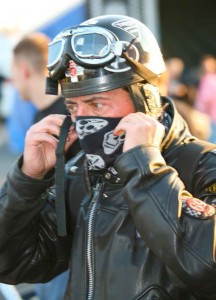 A biker wearing a vintage 'skid lid' helmet pulls a scarf over his mouth and nose before securing the strap of the helmet.