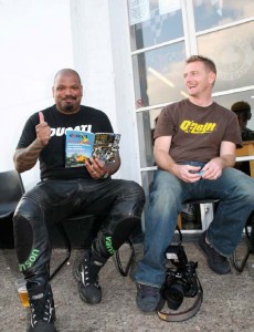 Two guys sitting outside on stools in front of the Ace front door, one wearing a Ducati t-shirt and leather bike jeans, giving a thumbs up while reading a copy of th10th anniversary Rider's Digest.