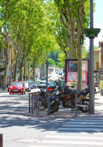 The KTM parked on a pavement, with some mopeds, in a tree lined street, in bright sunlight.