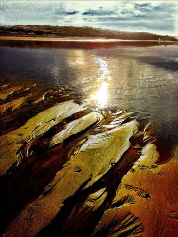 A beach at sunset, with the saturated sand just emerging from the water, lit by the sun's glow.